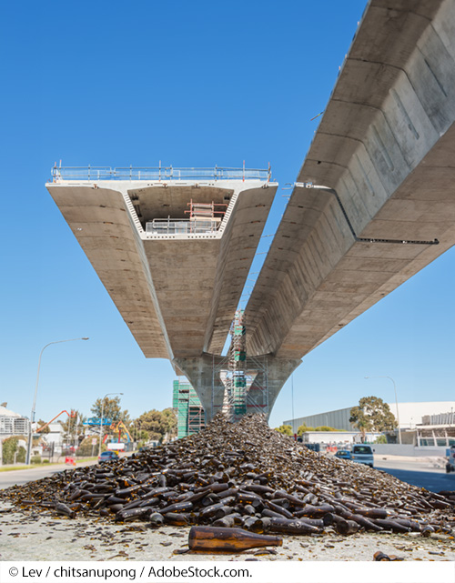 View of the underside of a bridge under construction with one lane completed and the other still in progress. Scaffolding, vehicles, and a large warehouse can be seen in the background, with a large pile of glass bottles laying underneath the bridge in the foreground. Image Source: © Lev / chitsanupong / AdobeStock.com.