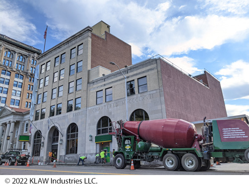 A concrete truck and workers pour concrete outside a building. In the background is a large bank. Image Source: © 2022 KLAW Industries LLC.