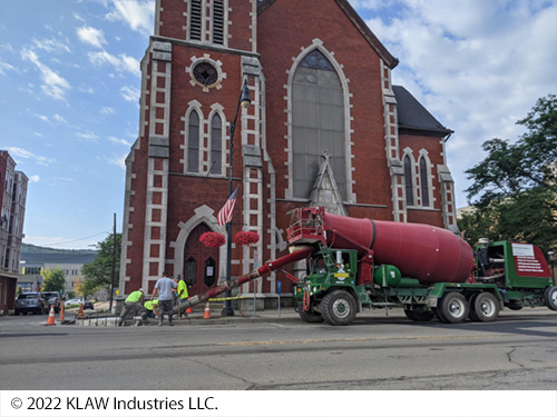 Workers move cement out of a cement mixer onto the roadway. A tall building is in the background. Image Source: © 2022 KLAW Industries LLC.