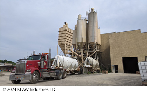 A large tractor-trailer and a ready-mix concrete plant in the background. Image Source: © 2024 KLAW Industries LLC.