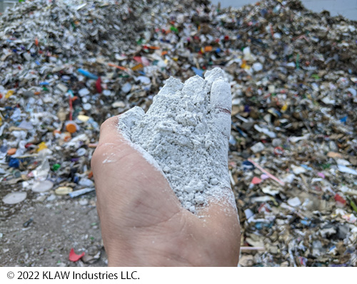 A hand holding SCM which looks like a pile of fine ash. In the background is a pile of waste glass. Image Source: © 2022 KLAW Industries LLC.