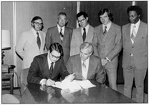 FHWA Administrator Norbert Tiemann (right) and UMTA Administrator Frank C. Herringer sign agreement on meeting urban transportation training needs. In the background, from left to right, Roger L. Dean, University and Industry Programs Officer, NHI; Roy W. Tidwell, NHI Director; Charles T Morrison, Jr., Training Coordinator, Office of Transit Management (OTM), UMTA; Stephen G. McConahey, Director of OTM, UMTA; and Dr. Frank E. Enty, Program Manager, Human Resources and Technical DeveloDment Division, OTM, UMTA.