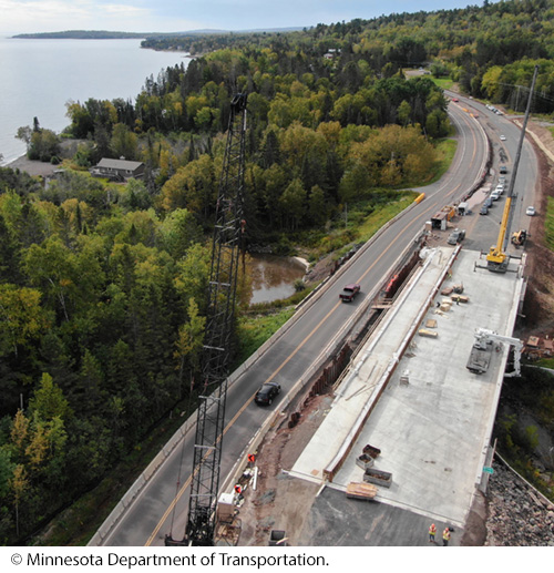  Aeriel view of bridge construction on a part of a multi-lane roadway lined with trees against a body of water. Image © Minnesota Department of Transportation.