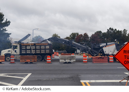 Two construction vehicles, including a dump trunk, between roadway construction safety barriers, barricades, and signs. Image © KirKam / AdobeStock.com.