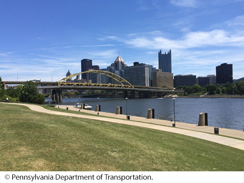 Side view of a bridge with vehicles over a body of water in front of a city’s daytime skyline. Image © Pennsylvania Department of Transportation.