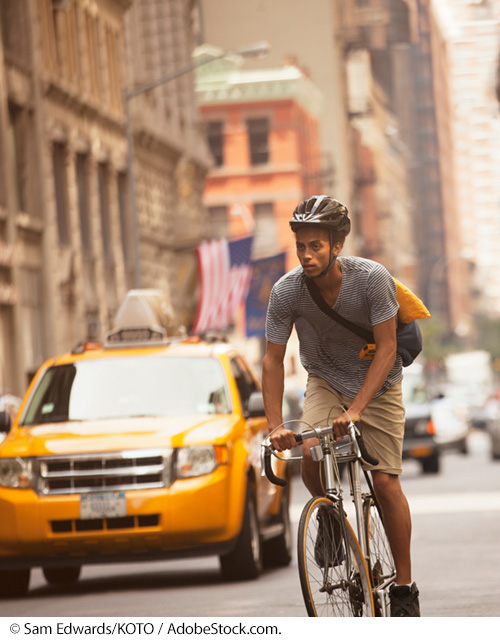 A bicyclist wearing a helmet rides on a crowded city street. High-rise buildings, taxis, and other vehicles can be seen behind the rider. Image Source: © Sam Edwards/KOTO / AdobeStock.com.