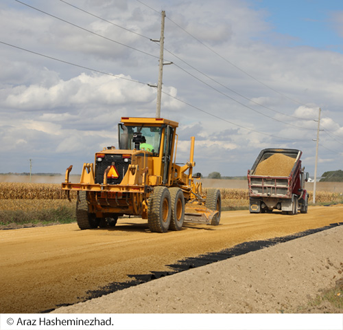 A road grader and dump truck spreading material over a roadway. Image Source: © Araz Hasheminezhad.