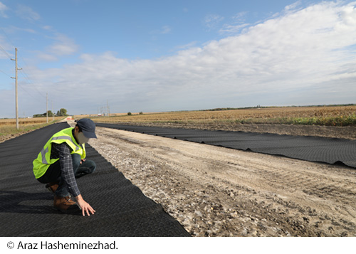 A worker in a safety vest bending down to touch a black paved section of roadway. Image Source: © Araz Hasheminezhad.