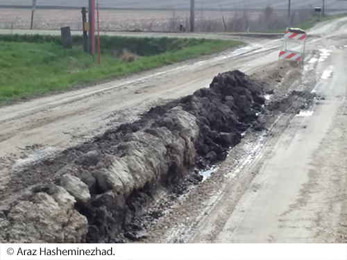 A rural, dirt road with ruts along its length. The middle of the road contains plowed snow in a trench. A traffic barricade is at the end of the trench. Image Source: © Araz Hasheminezhad.