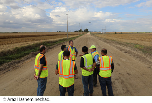 A group of people in safety vests standing on a rural dirt road. A drone flies in the distance. Image Source: © Araz Hasheminezhad.