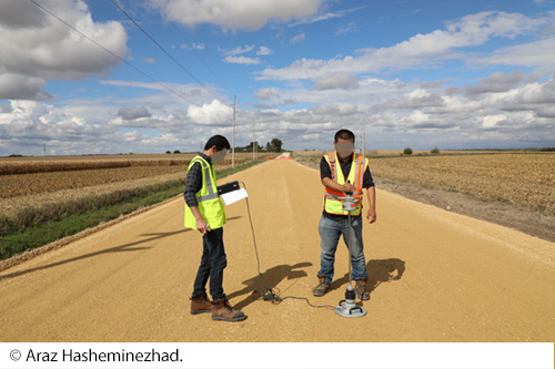 Two workers in safety vests using a measuring device to take readings on a rural dirt roadway. Image Source: © Araz Hasheminezhad.