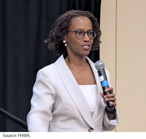 An African-American woman holds a microphone in front of a black cloth backdrop. Image Source: FHWA.