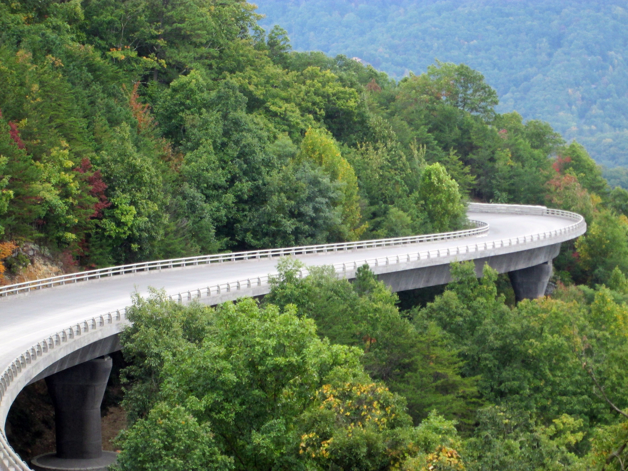 A winding road with a bridge-like structure, surrounded by lush green trees. The landscape features a mix of trees with some showing signs of autumn colors. The road curves through the mountainous area with a clear blue sky in the background.