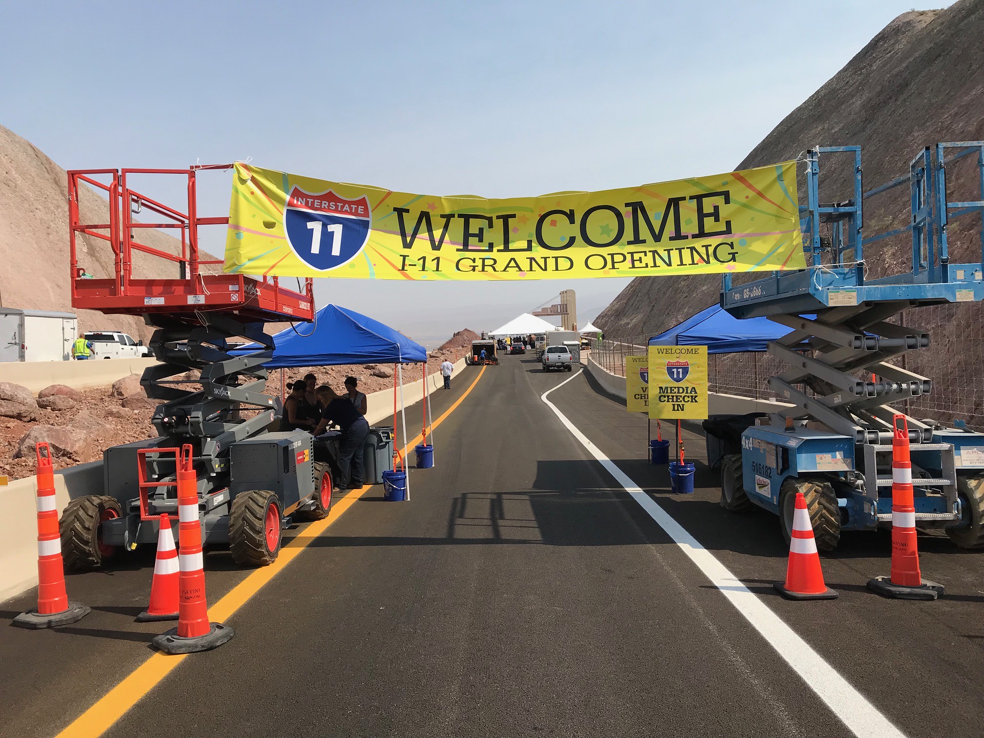 A grand opening event for Interstate 11, with a large banner reading "WELCOME I-11 GRAND OPENING" across the road. There are people gathered under tents for media check-in, and a construction vehicle is in the foreground. The surrounding terrain is rocky, and the road looks newly paved.
