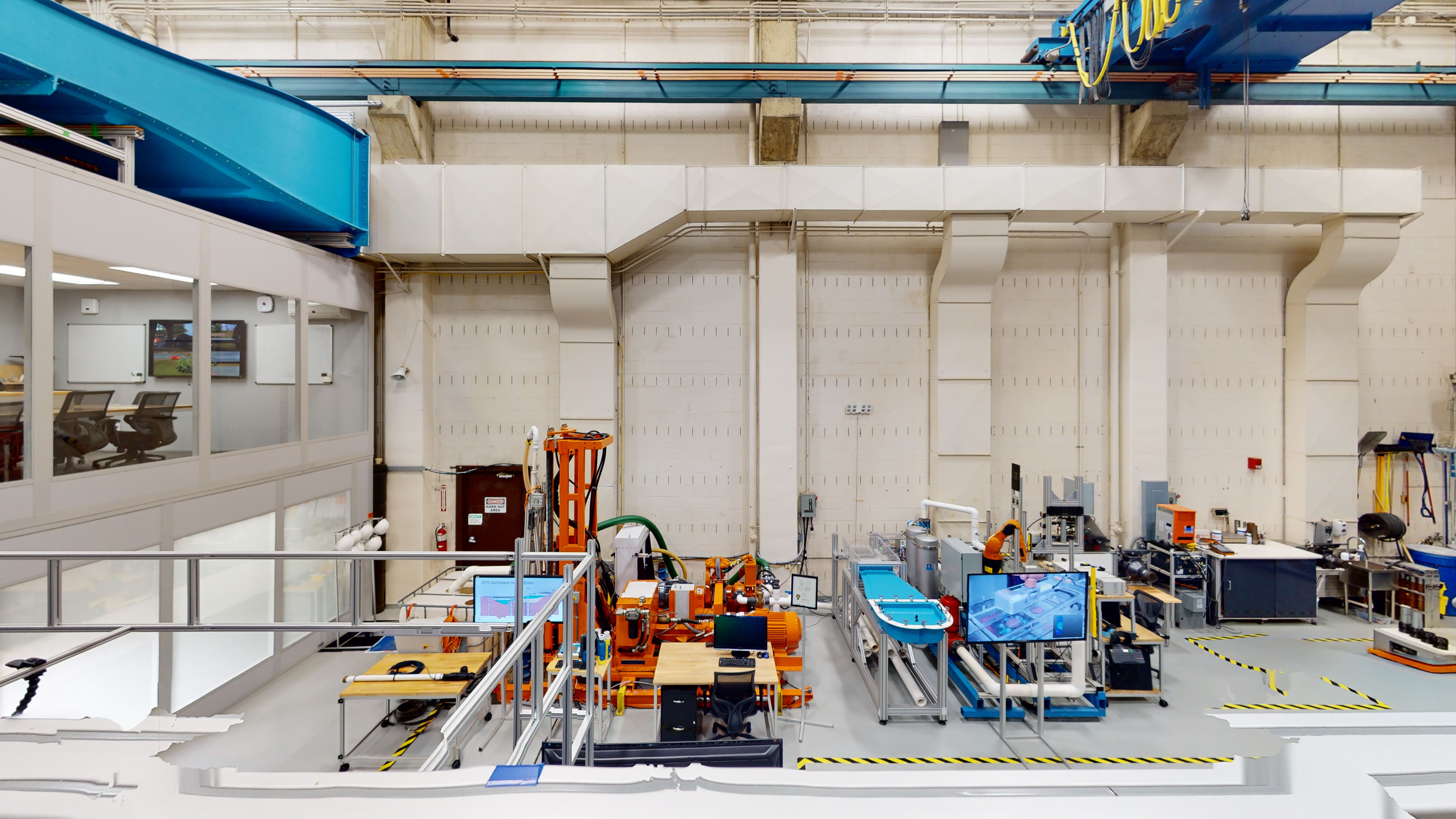 Overview shot of equipment in the Hydraulic Laboratory, including an in-situ scour testing device (ISTD), a drill bit in a soil chamber, computer workstations, a basin with several clamps that lead to a solid portion and clear flow channel, part of the ex-situ scour testing device (ESTD) station (which features a robotic arm), stand-mounted monitors, and a soil compaction station (which features a hydraulic press). 