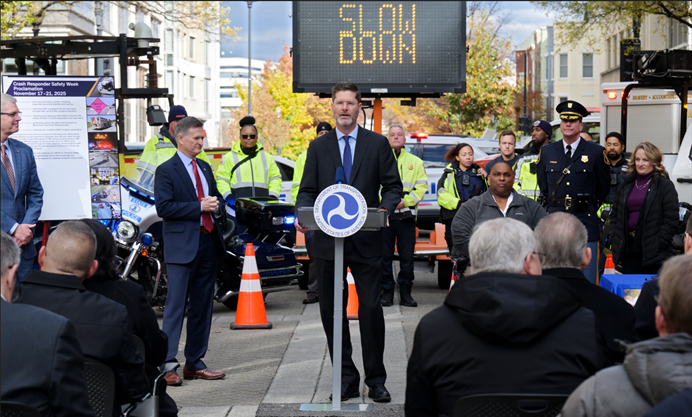 Officials presenting at a USDOT event with a focus on transportation safety. A poster board and various law enforcement motorcycles are visible in the background. FHWA Administrator McMaster speaking at the podium.