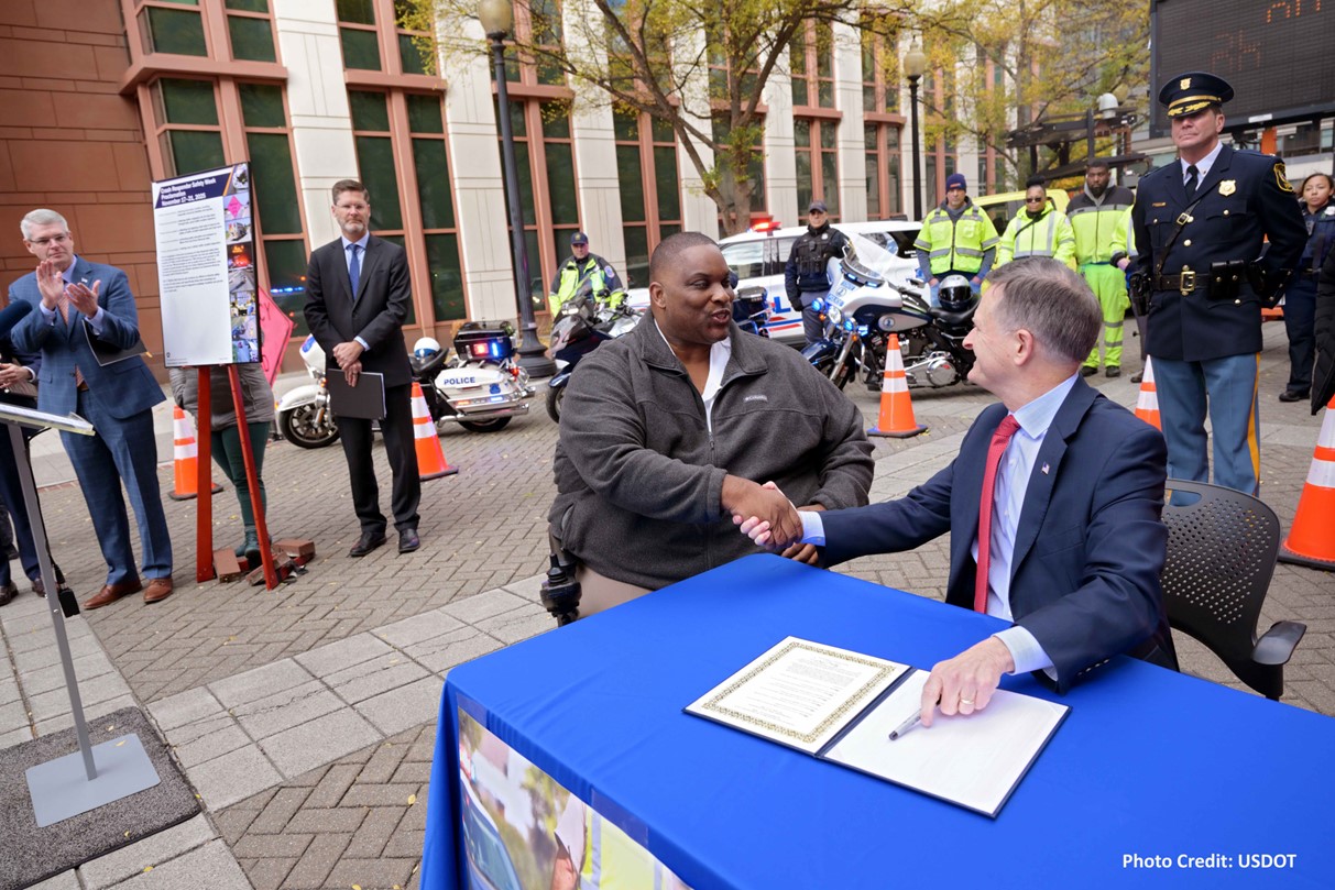 Two individuals shaking hands at an outdoor event with spectators and police officers in the background. A folder is open with a document on a table for signing, and there are motorcycles parked nearby. Photo credit: USDOT.