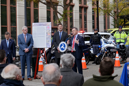 Officials presenting at a USDOT event with a focus on transportation safety. A poster board and various law enforcement motorcycles are visible in the background. USDOT Deputy Secretary Bradbury speaking at the podium.