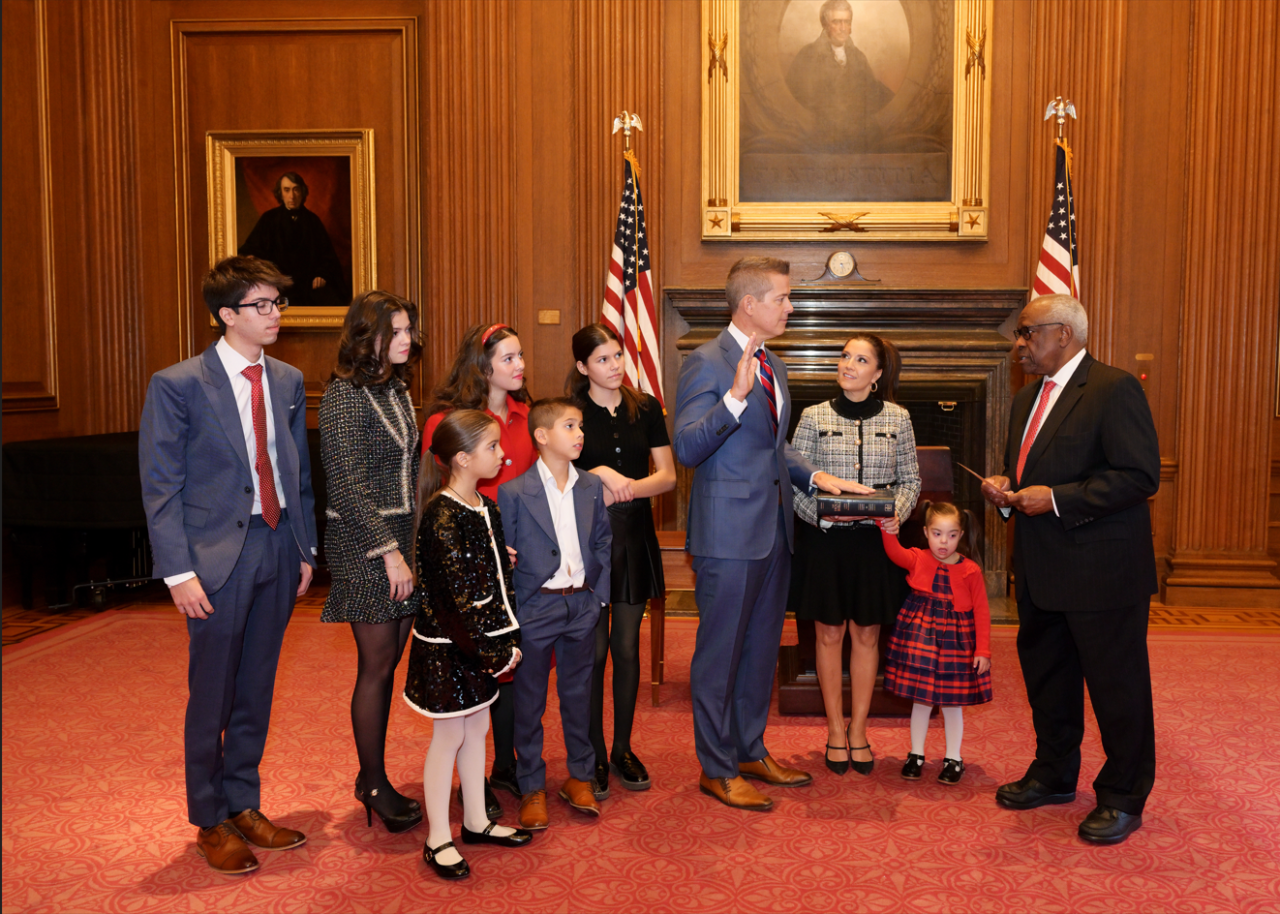 Sean Duffy stands in a room in front of a fireplace flanked by two U.S. flags, raising his right hand with his left hand on the Bible, beside his wife, with six of his children standing beside him and one child standing next to his wife, with Justice Clarence Thomas standing opposite them.