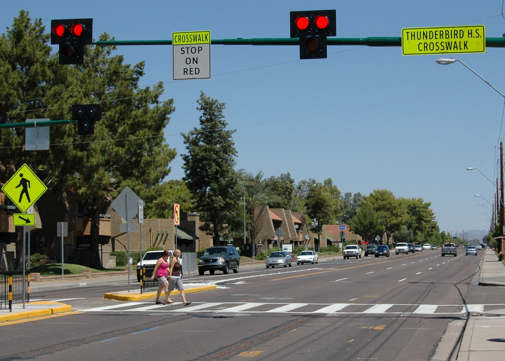 five-lane road running through a suburban town with two women crossing the street at a crosswalk with a Pedestrian Hybrid Beacon while cars are approaching