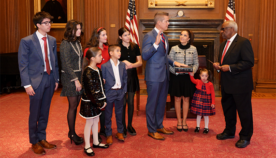 Sean Duffy stands in a room in front of a fireplace flanked by two U.S. flags, raising his right hand with his left hand on the Bible, beside his wife, with six of his children standing beside him and one child standing next to his wife, with Justice Clarence Thomas standing opposite them.