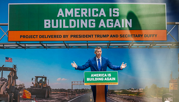 DOT Secretary Sean Duffy standing at a podium in front of an image of road construction with a simulated traffic sign "America is Building Again"