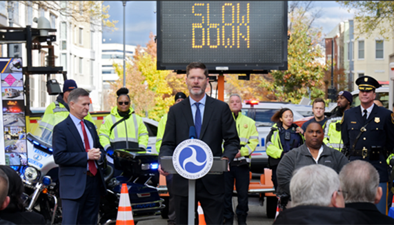 FHWA Administrator McMaster speaking at the podium during USDOT crash responder safety event. A poster board and various law enforcement motorcycles are visible in the background.